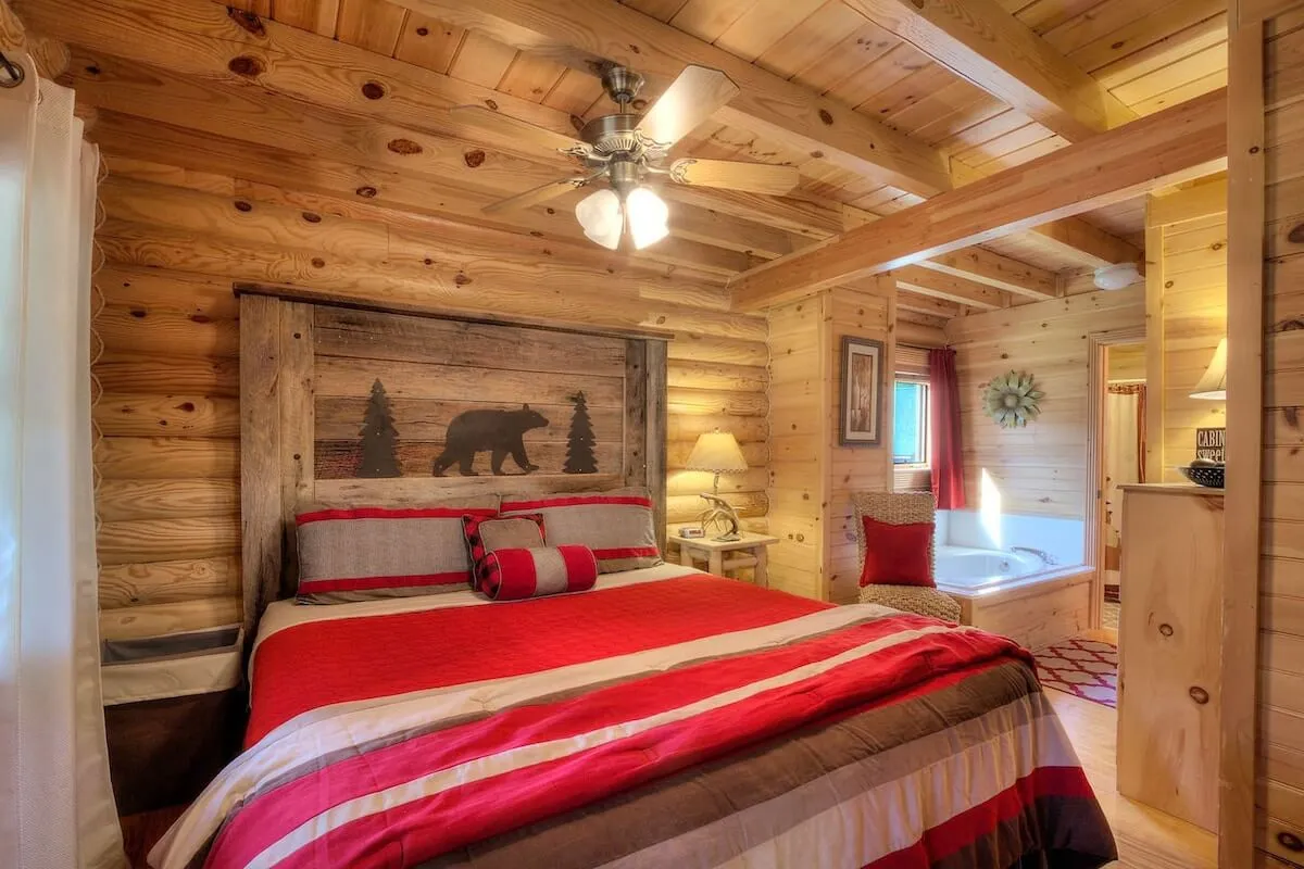 The image showcases a cozy bedroom interior with a bear-themed headboard, inviting striped bedding, rustic wooden beams, and a soothing bathtub. The scene exudes warmth and comfort, suggesting opportunities for relaxation, such as unwinding in bed or enjoying a candlelit bath while surrounded by natural wood accents.