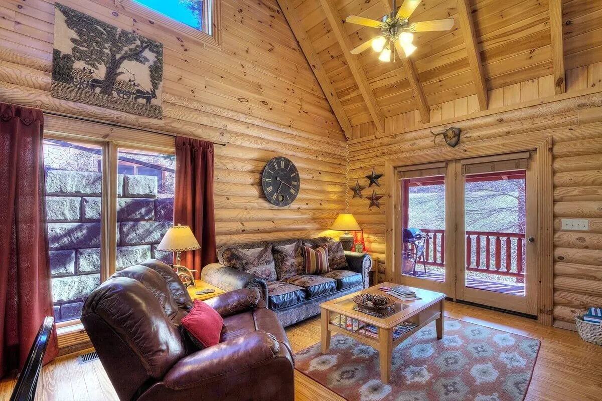 The living room features a plush leather sofa and a wooden coffee table, bathed in natural light from large windows that offer tranquil wooded views. Warm decorative touches and a table lamp contribute to a cozy, inviting atmosphere ideal for both socializing and quiet relaxation.