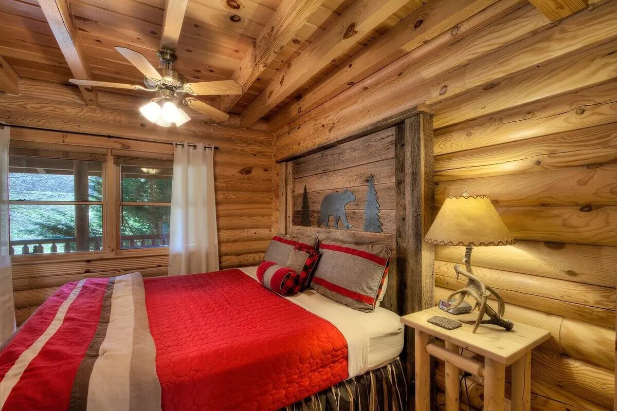 A warm, inviting bedroom with a comfortable bed adorned in a red quilt, wooden headboard, cozy lamp-lit nightstand, and a window with curtains that let in natural light and offer a glimpse of the peaceful outdoors.