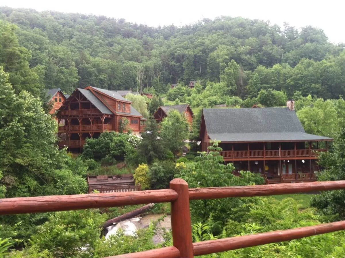 A picturesque exterior view of several log cabins nestled among lush greenery, with a wooden railing and a dramatic mountain backdrop. The scene exudes tranquility and togetherness, suggesting a perfect base for family gatherings and outdoor fun.