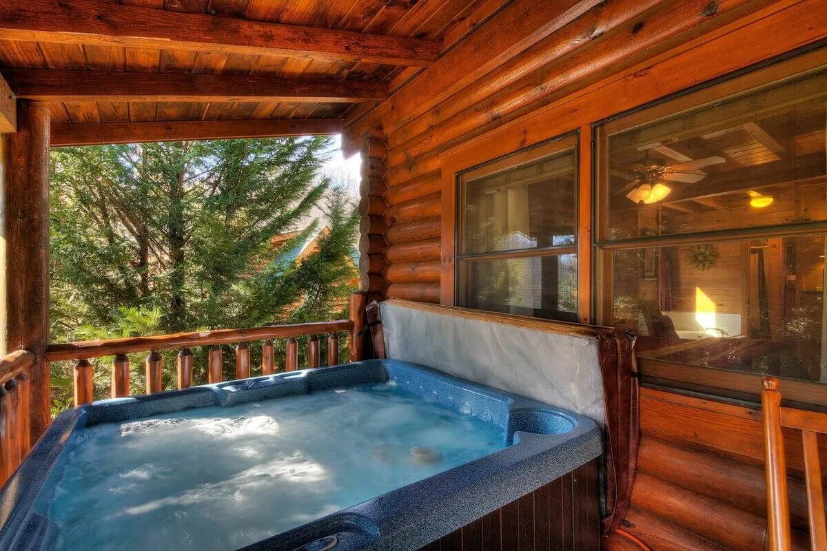 This image showcases a tranquil outdoor deck area featuring a bubbling hot tub with a wooden railing and surrounded by towering trees. The peaceful forest setting and the inviting water in the hot tub immediately evoke a sense of relaxation and connection with nature.