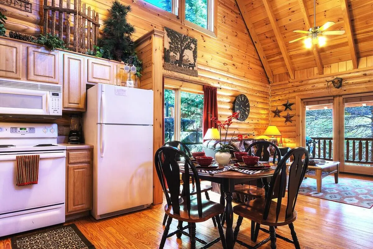 A warmly lit open-concept kitchen and dining area featuring modern appliances, a wooden dining table set for a meal, expansive windows letting in natural light, exposed wooden beams, and decorative plants. The kitchen counters are spacious and inviting, seamlessly connecting to the dining area where people can easily gather.