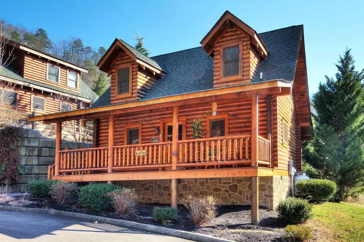 A charming log cabin nestled amidst landscaped gardens. The front porch, with its inviting seating, faces the sunset and is surrounded by lush greenery. The gabled roof and warm wooden exterior create a cozy first impression, and the tranquil atmosphere is enhanced by the natural surroundings.