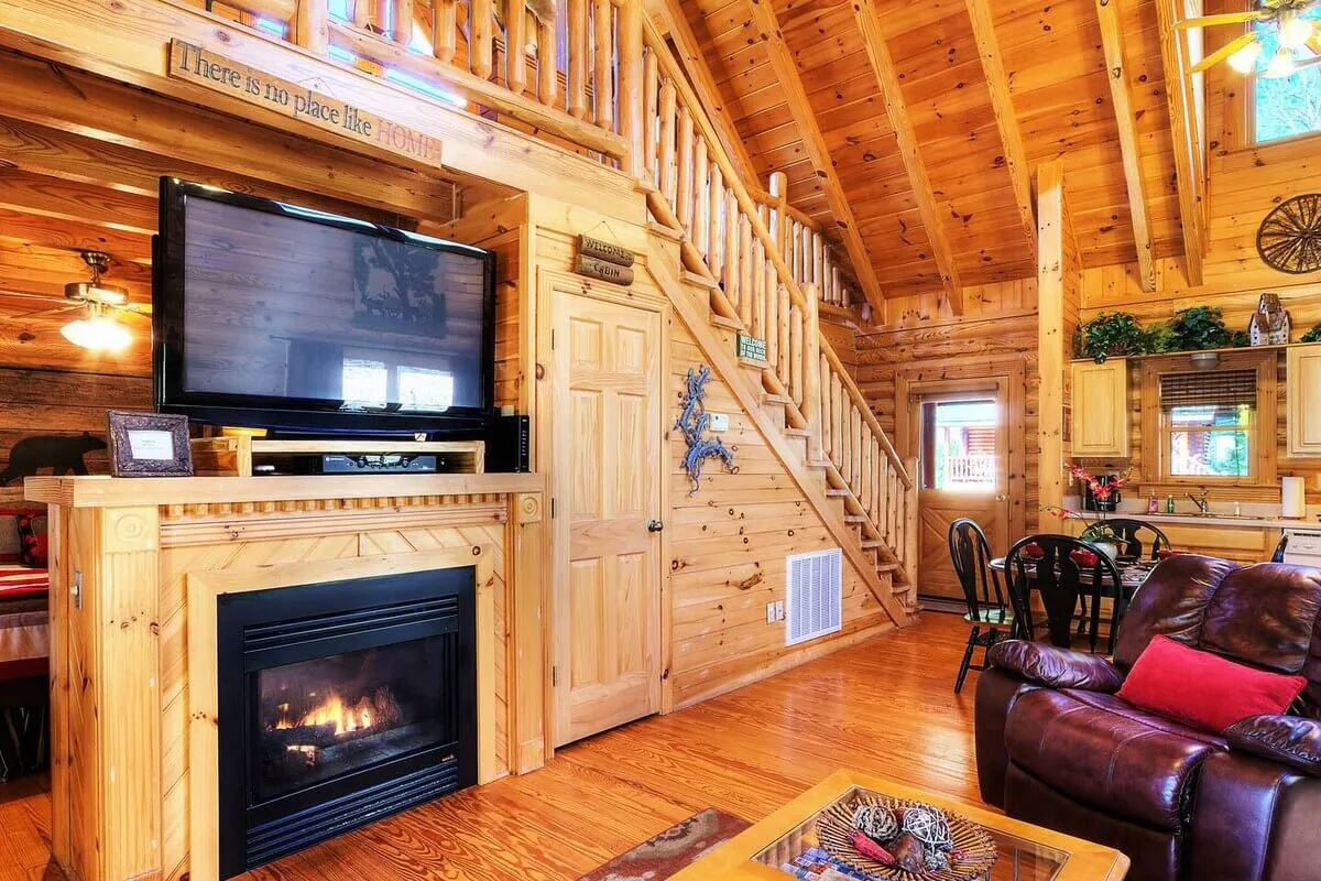 A warm and spacious living area showcasing a stone fireplace, adjacent television, and a staircase leading to a loft. The scene features wooden walls and a glimpse of a nearby dining area, all arranged for group gatherings and intimate evenings by the fire.