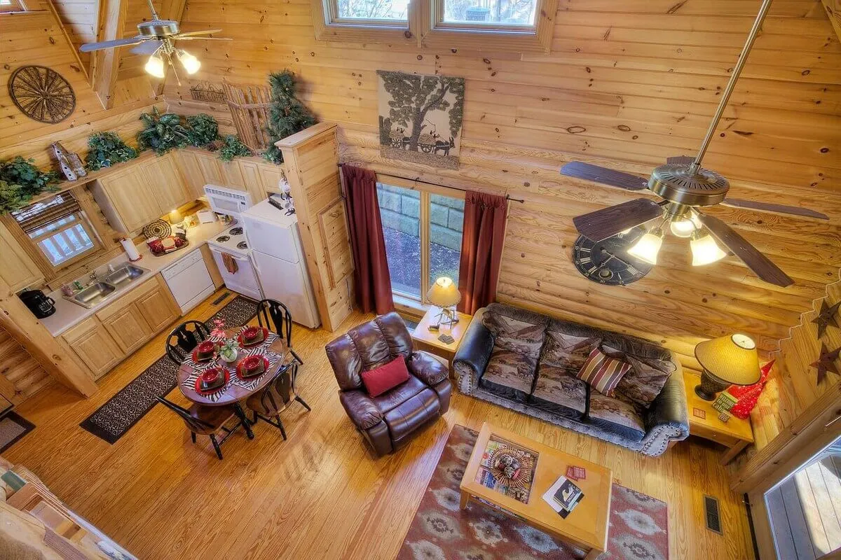 A wide-angle view of the open-plan living space, blending a comfortable seating area, dining table, and kitchen. Large windows flood the room with natural light, while the wooden ceiling and décor maintain a cozy cabin ambiance. The scene conveys togetherness and connection.