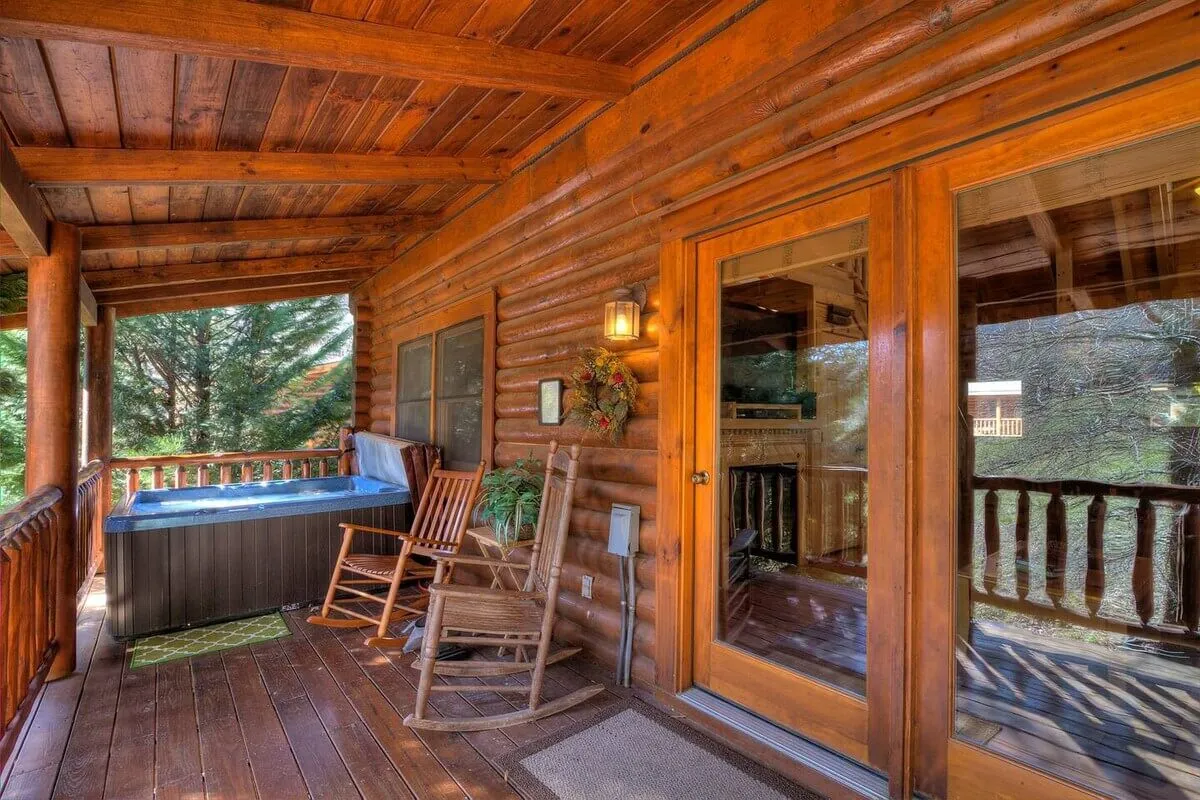 The image presents a cozy deck space with a hot tub, a rocking chair, wooden railings, and a decorative wreath on the door. The arrangement is inviting, blending comfort with rustic charm, and highlights both the hot tub and the option to relax on the deck seating.