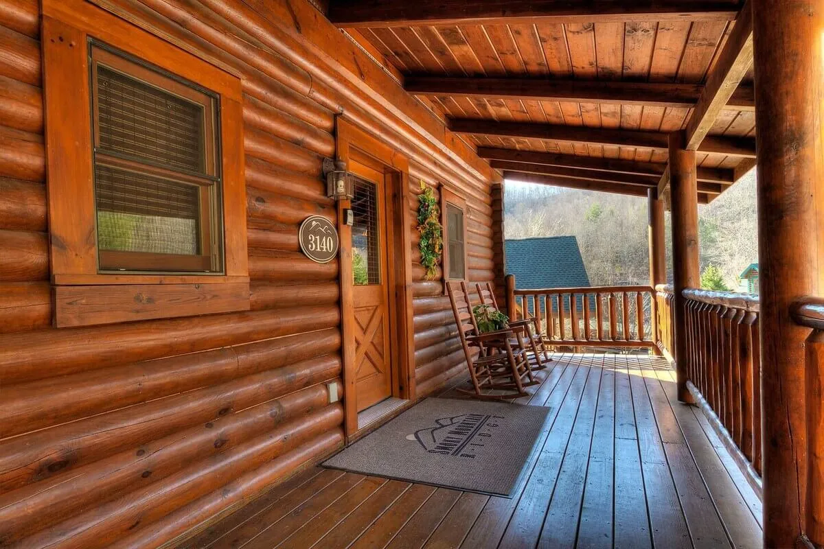 This image features the inviting log cabin exterior, complete with wooden rocking chairs on the porch and a welcoming front door adorned with a lantern and a house number sign. The lush greenery and mountainous backdrop evoke a sense of seclusion and connection to nature.