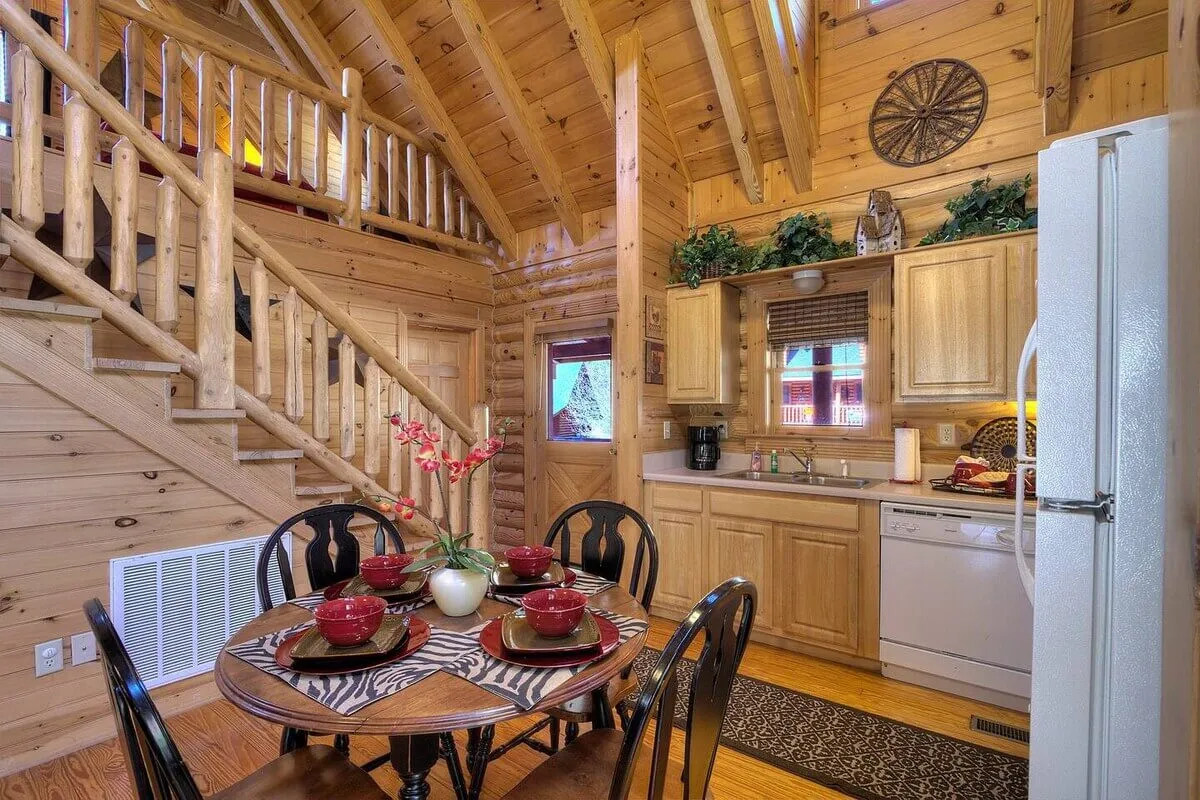 A cozy, inviting dining area with a table set for four, adjacent to a well-equipped kitchen with visible appliances. The area is warmly illuminated, with a staircase leading up to a loft, and the layout encourages sociable interactions and lingering conversations.
