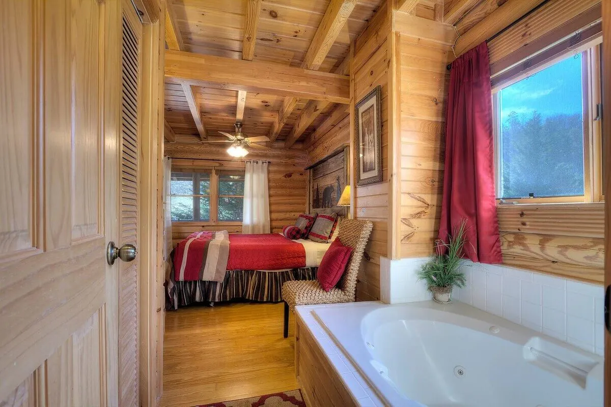A spacious bedroom bathed in natural light, featuring a king-sized bed with plush red bedding, a whirlpool tub conveniently located in the room, and large windows offering tranquil woodland views. The wooden beams and walls add warmth and a rustic, cozy atmosphere.