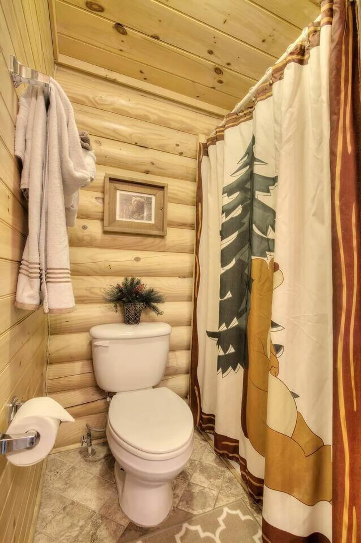 A well-appointed bathroom with rich wooden walls, a decorative shower curtain featuring woodland motifs, soft towels on a rack, a framed picture, and a small plant, exuding warmth and comfort.